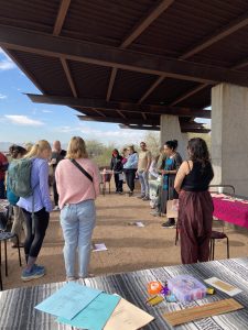 Group of people gathered under a shaded pavilion at an outdoor craft fair; tables with colorful papers and supplies in the foreground.