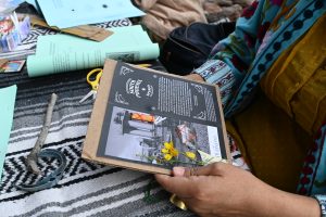 Person sits outdoors, holding a booklet titled 'South Phoenix Speaks' amid craft materials like scissors, flowers, and papers on a striped blanket, engaged in a project.