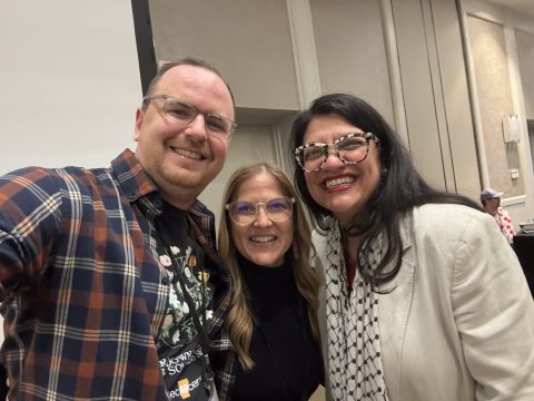 Three people taking a close-up group selfie indoors, smiling at the camera (man left in plaid shirt, two women center and right).