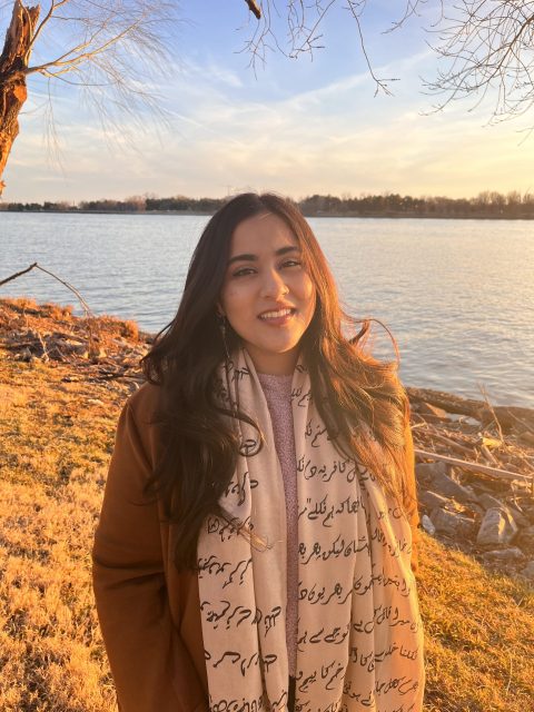 Woman standing near a lake at sunset, wearing a scarf with script and a brown coat, smiling at the camera.