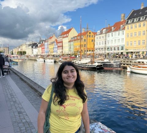 Smiling woman in a yellow shirt stands by a canal with colorful row houses and boats in the water behind her.
