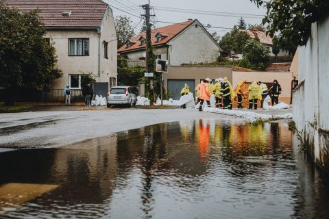 Flooded residential street with workers in yellow rain ponchos placing sandbags along the curb.