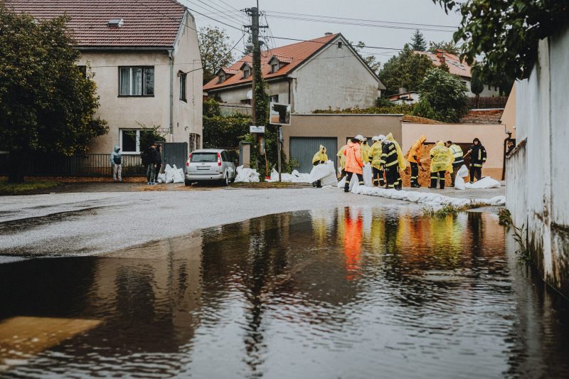 Flooded residential street with workers in yellow rain ponchos placing sandbags along the curb.