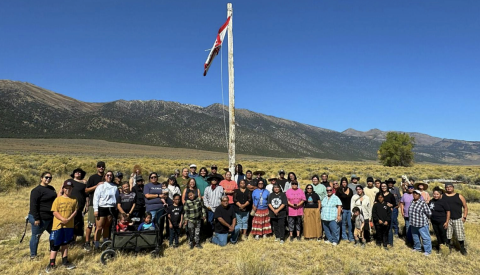 Diverse group of people posing for a photo in a grassy field with a tall flagpole and distant mountains under a blue sky.