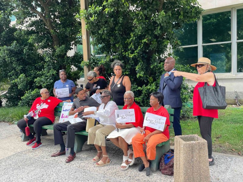 Group of adults sitting on a green bench outdoors, holding signs about lowering utility rates at a community gathering.
