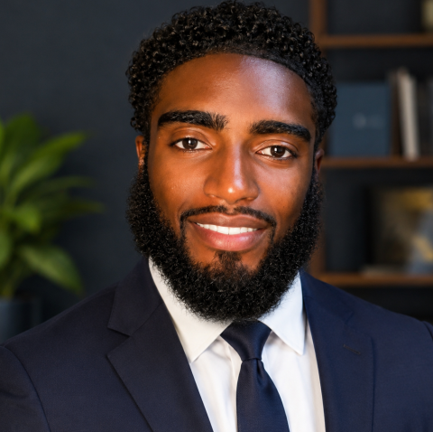 Portrait of a smiling man with a full beard in a navy suit and white shirt indoors in a professional setting.