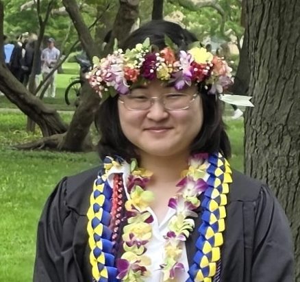 Graduate wearing a flower crown and lei, smiling outdoors in cap and gown.