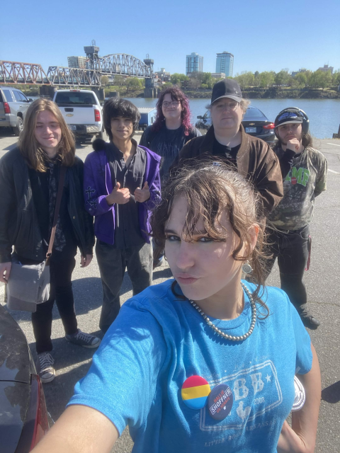 Five friends posing for a selfie by a river, with a steel bridge and city buildings in the background.